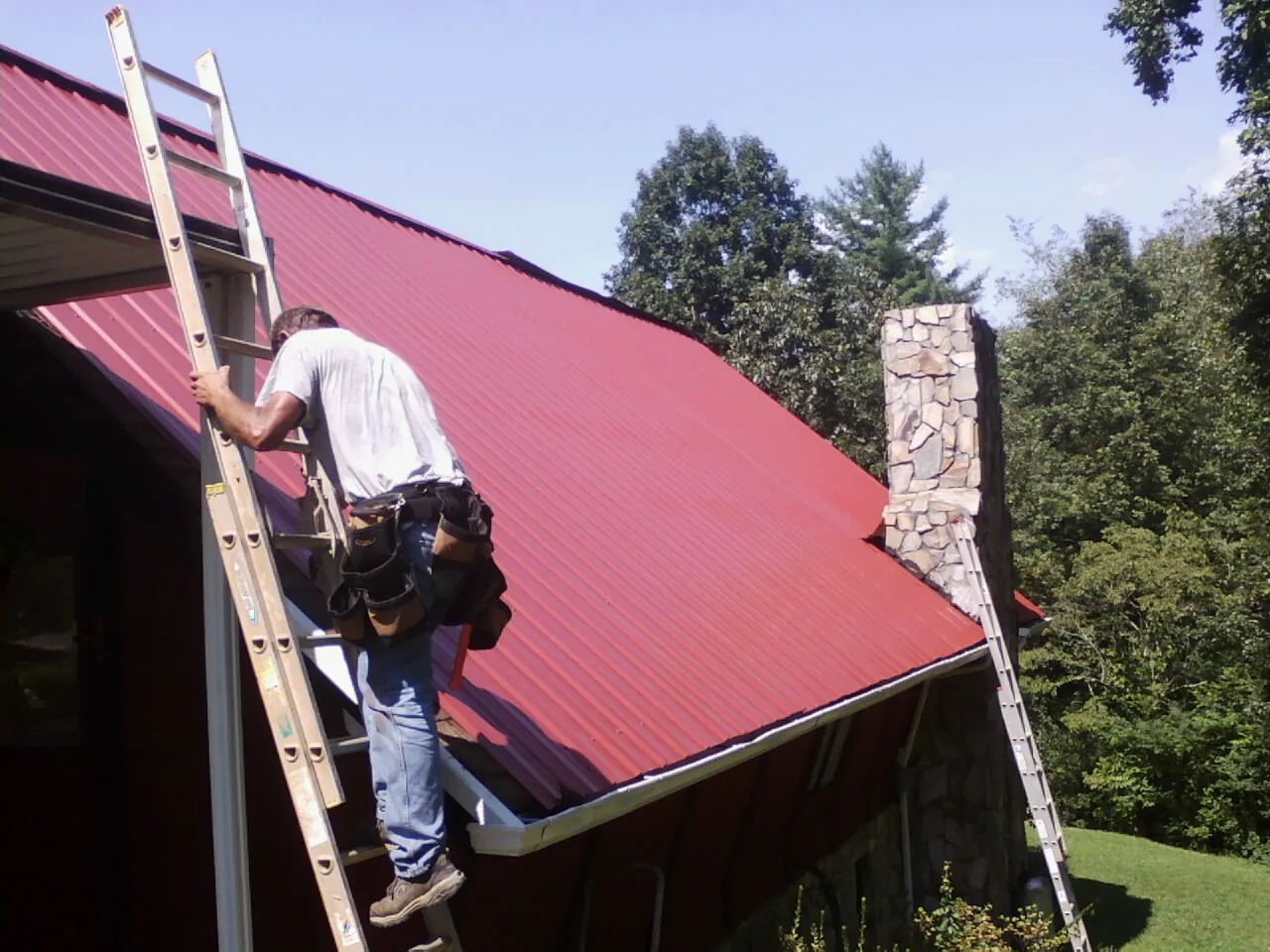 Roofer on a ladder beside a red metal roof and stone chimney — Soffit & Fascia Repair by Roofing Brothers in Kanawha